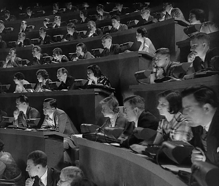 Still from The Testament of Dr. Mabuse. Seven rows of desks are visible in a lecture hall, every seat occupied by a person listening intently.