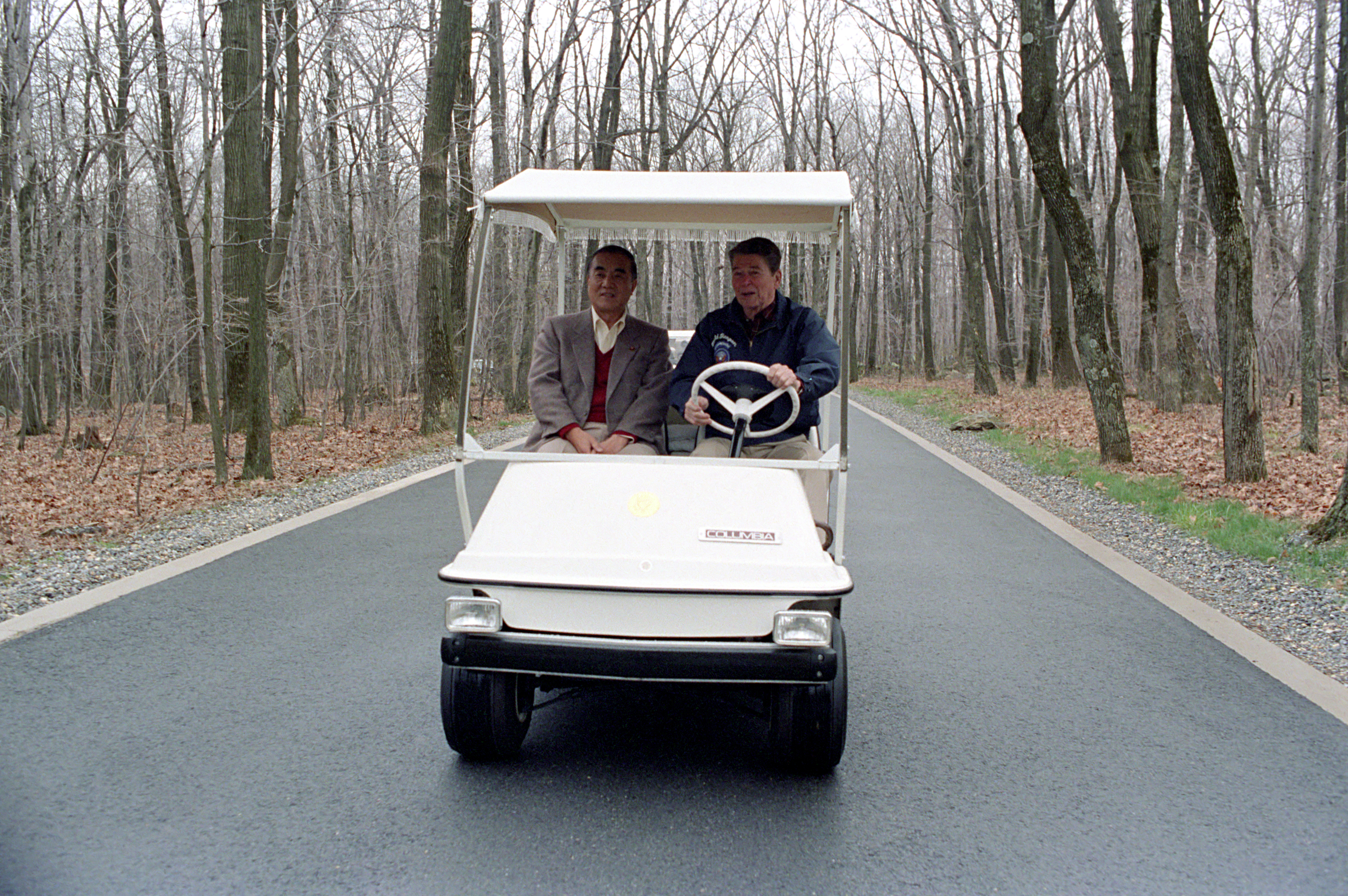 Photograph of Ronald Reagan and Nakasone Yasuhiro in a golf cart. Reagan is driving. It is April, and there are no leaves on any trees in the foggy woods around them.