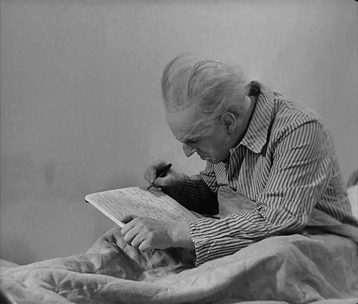 Still from The Testament of Dr. Mabuse. Against a blank, white wall, Dr. Mabuse sits in a bed, sheets up to his chest, hunched over a notebook in which he feverishly writes something with a pen. His shirt is striped, his gray hair thin and unkempt. He is viewed in profile, emphasizing the pointed nose.