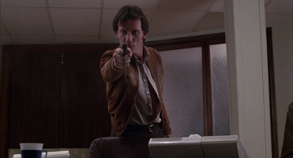 Screenshot from Videodrome. Max aiming a gun at the camera, killing his former colleagues.