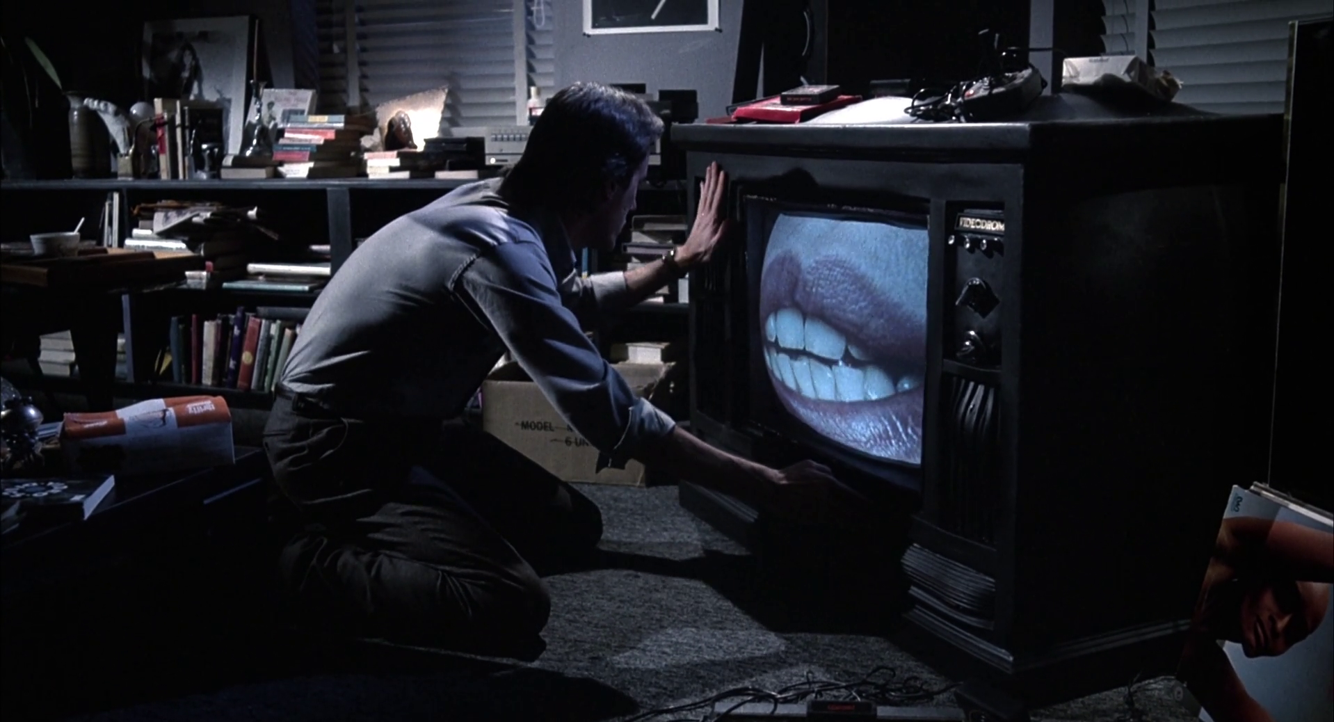 Still from Videodrome. In a dimly lit apartment, Max Kenn kneels in front of a throbbing TV set that he caresses. On the screen is an extreme close-up of a woman's lips speaking to him.
