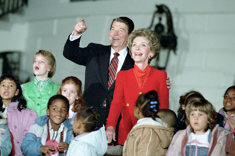 Standing over a group of children with various expressions, Ronald Reagan has one arm around Nancy Reagan and points to something out of frame. Both he and Nancy are grinning, and Nancy looks almost entranced.