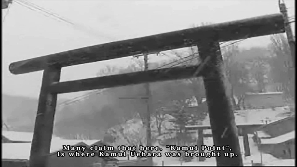Screenshot from "LifeCut." A black-and-white still of video showing a shinmei torii in a snow-covered area. There are trees, powerlines, and several buildings visible behind the torii closest to the camera. Subtitles read, "Many claim that here, 'Kamui Point', is where Kamui Uehara was brought up."