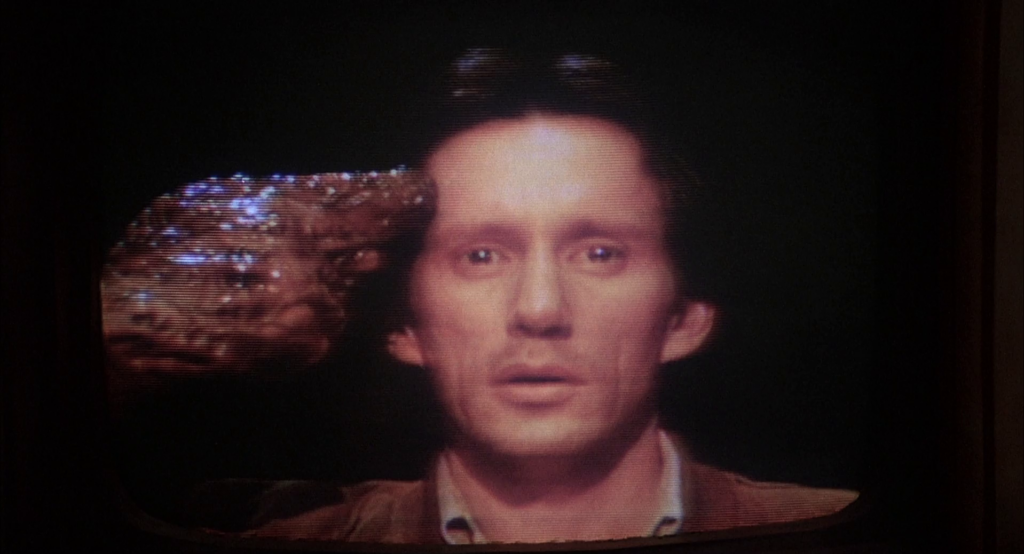 Still from Videodrome. On a TV set, Max holds a gun to his head. His expression suggests a mystical experience. The gun is sealed to his hand, both of which are overgrown with an additional layer of shiny, brown flesh.